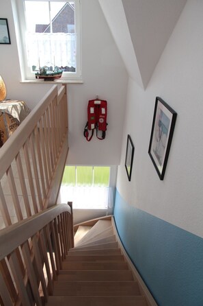 Una escalera con barandillas de madera que conduce a una plataforma. Un pequeño modelo de barco está colocado en un alféizar, y un extintor está fijado a la pared.