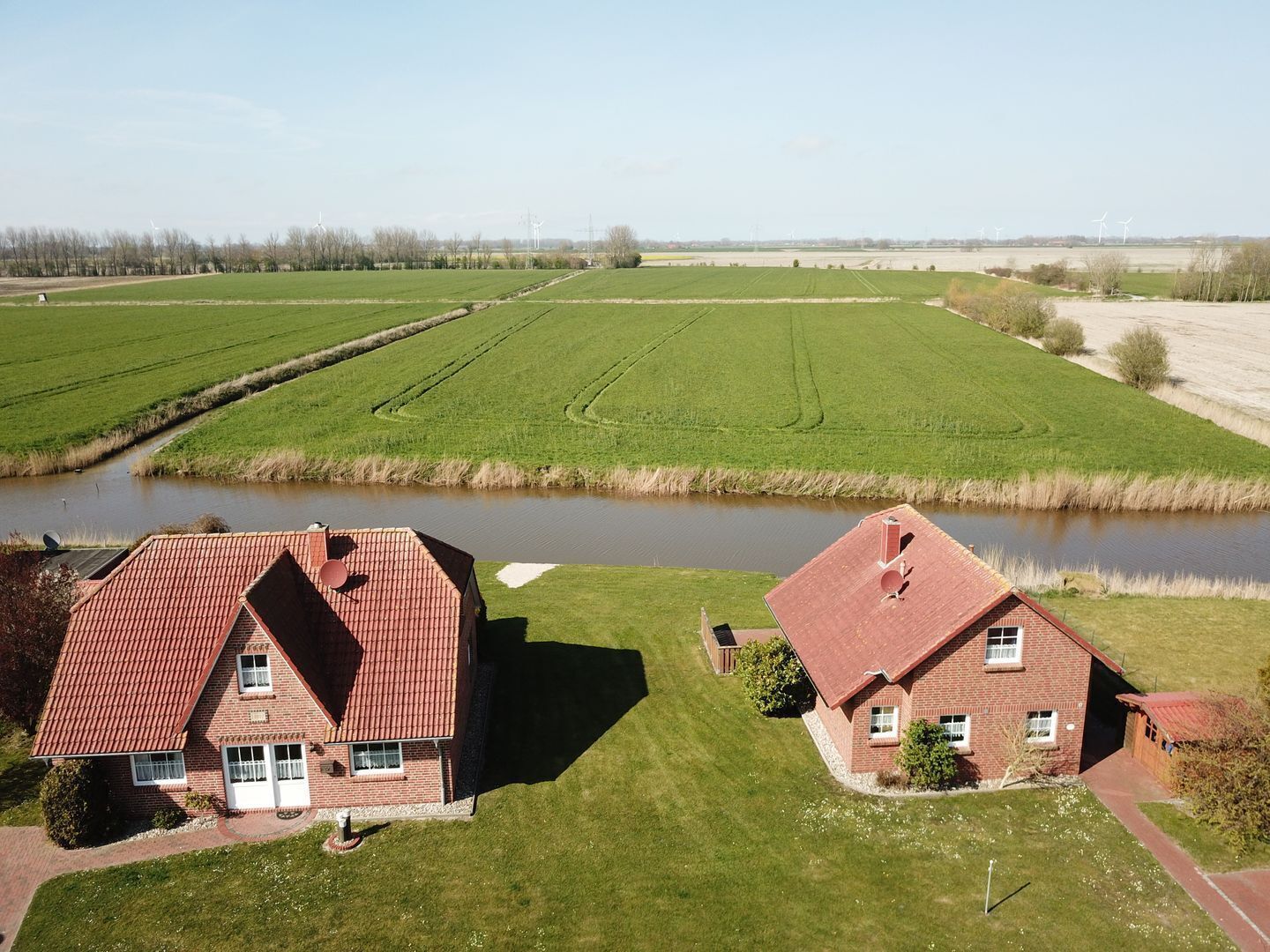 Dos casas de ladrillo con techo rojo separadas por un río, rodeadas de campos verdes y turbinas eólicas a lo lejos.