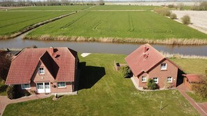 Deux maisons en briques aux toits rouges, séparées par une rivière, entourées de champs verdoyants et, au loin, on aperçoit des éoliennes.