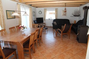 A cozy living room with a wooden dining table, chairs, a black sofa, a TV, and a wood-burning stove. - Wiesenblick Fenja - Wiesenblick Fenja Vacation Home: Sauna, Rowing Boat & Water View (Krummhörn)