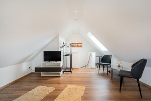 A modern, well-lit attic room with a wooden floor, a TV on a stand, a desk with a chair, and a small table with a lamp. - Leuchtfeuer - Leuchtfeuer Vacation Rental with a Balcony in Greetsiel (Krummhörn)