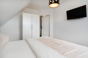 A bedroom with a bed, a wardrobe, a television mounted on the wall, and a ceiling light fixture. - Leuchtfeuer - Leuchtfeuer Vacation Rental with a Balcony in Greetsiel (Krummhörn)