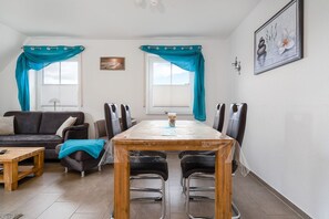 A room with a wooden dining table, four chairs, a sofa, and a coffee table. There are two windows with curtains, a painting on the wall, and a lamp. - Leuchtfeuer - Beautiful vacation apartment (75sqm) for 4 persons and dog with balcony in (Krummhörn)