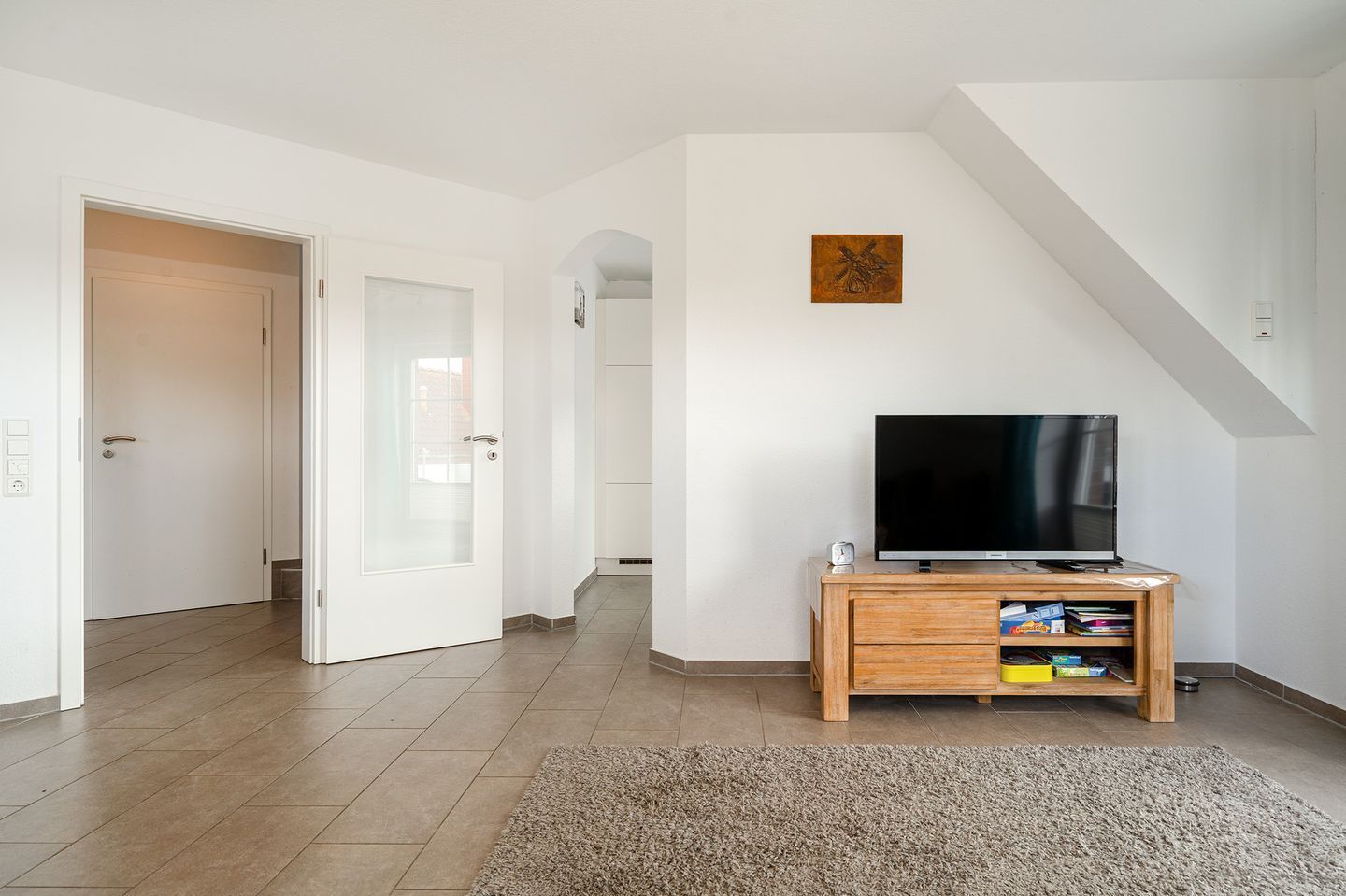A modern living room with a flat-screen TV on a wooden stand, a small rug, and a staircase to the right.