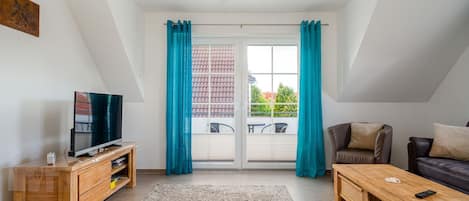 En moderne stue med fjernsyn, træmøbler, en sofa og udsigt til en balkon med blå gardiner.