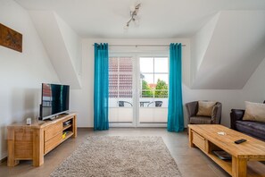 A modern living room with a TV, wooden furniture, a sofa, and a view of a balcony with blue curtains. - Leuchtfeuer - Leuchtfeuer Vacation Rental with a Balcony in Greetsiel (Krummhörn)