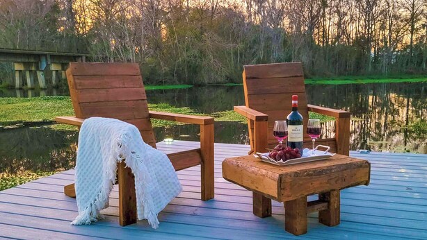 Zwei hölzerne Adirondack-Stühle auf einer Holzterrasse an einem ruhigen See, mit einer Weinflasche und Trauben auf einem kleinen Tisch.