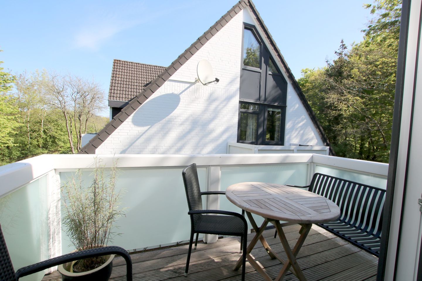 A balcony with a table and chairs, a potted plant, and a white building with a triangular roof and large windows.