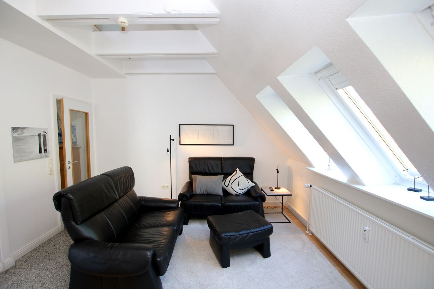 Une chambre de style loft moderne, avec un plafond en pente, un canapé en cuir noir, un pouf assorti et une œuvre d’art encadrée accrochée au mur.