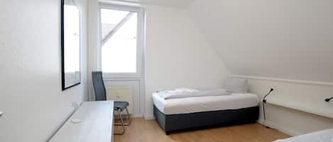 Um quarto pequeno e bem iluminado, com uma cama, uma escrivaninha e uma cadeira.