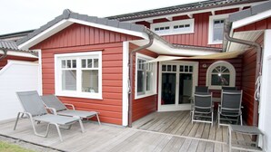 Une maison rouge avec une terrasse en bois, deux chaises longues et une table.