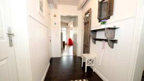 Un couloir étroit, doté d’un plancher sombre, de murs blancs et d’une porte blanche sur le côté gauche. On y trouve un petit banc blanc et une étagère ornée d’objets décoratifs, situés sur le côté droit.