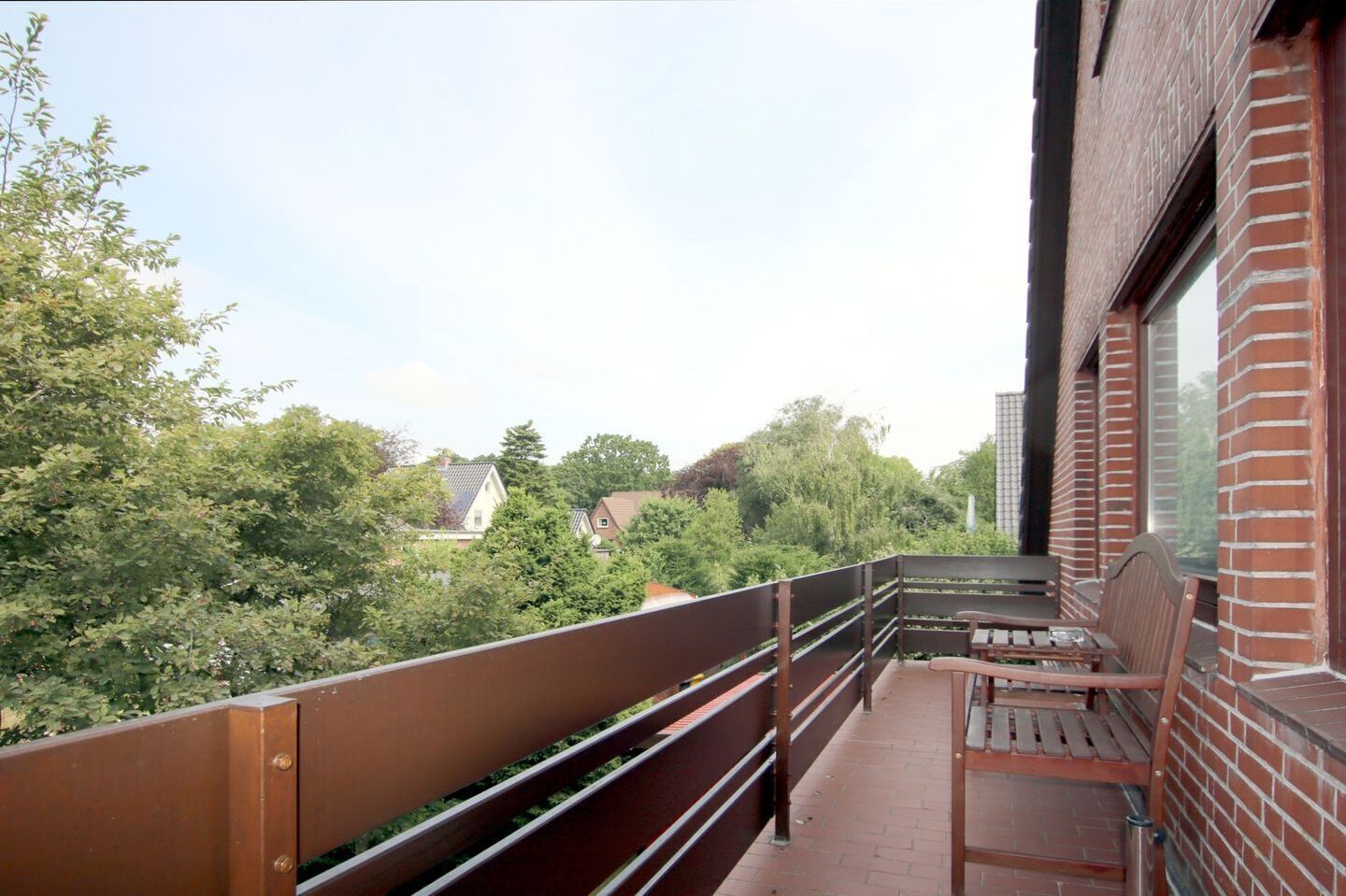 Un balcone con una panchina in legno e una ringhiera, affacciato su una zona residenziale con alberi e case.