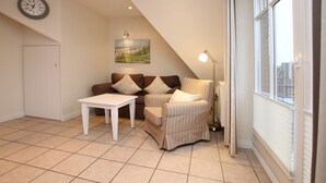 Een gezellige woonkamer met een bank, fauteuil en een kleine tafel. Er hangt een klok aan de muur, een schilderij en er is een raam met zonwering.