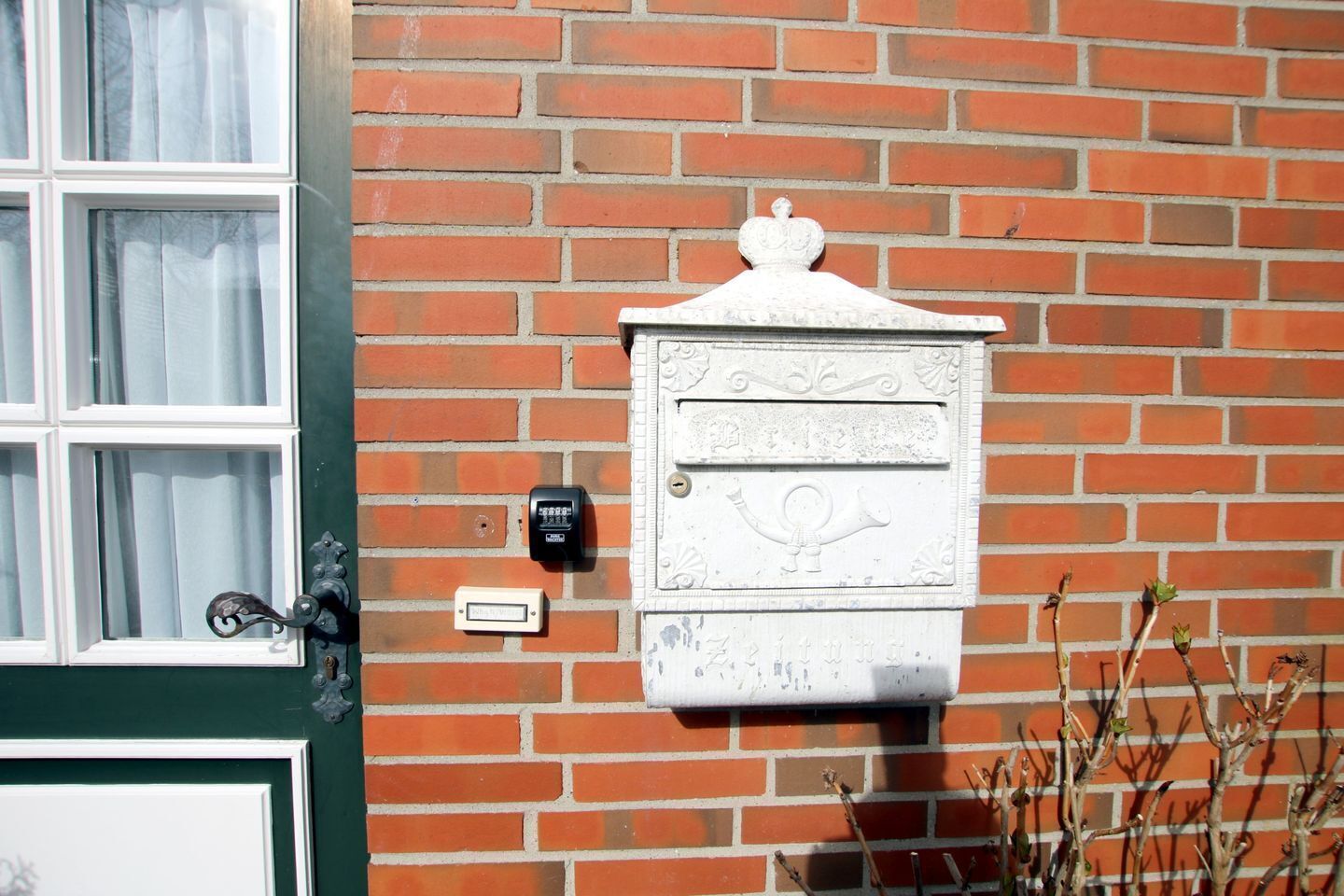 Une boîte aux lettres traditionnelle, ornée d’un élément décoratif sur le dessus, dotée d’une sonnette et d’une fenêtre avec des rideaux.