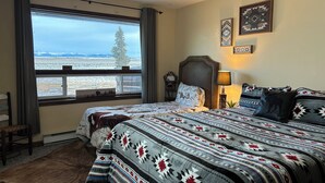 Un dormitorio con un ventanal grande, una cama con ropa de cama estampada, una mesita de noche, una lámpara y adornos en la pared.