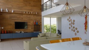 A modern living room with a large flat-screen TV, a sofa, a dining table with chairs, and a balcony with a view of greenery. - WONDERFUL HOUSE - HIGH WALL/ PORTO DE GALINHAS (IPojuca)