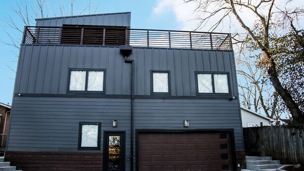Een modern gebouw met twee verdiepingen, een donkere gevel, een oprit en een klein balkon op de bovenste verdieping.