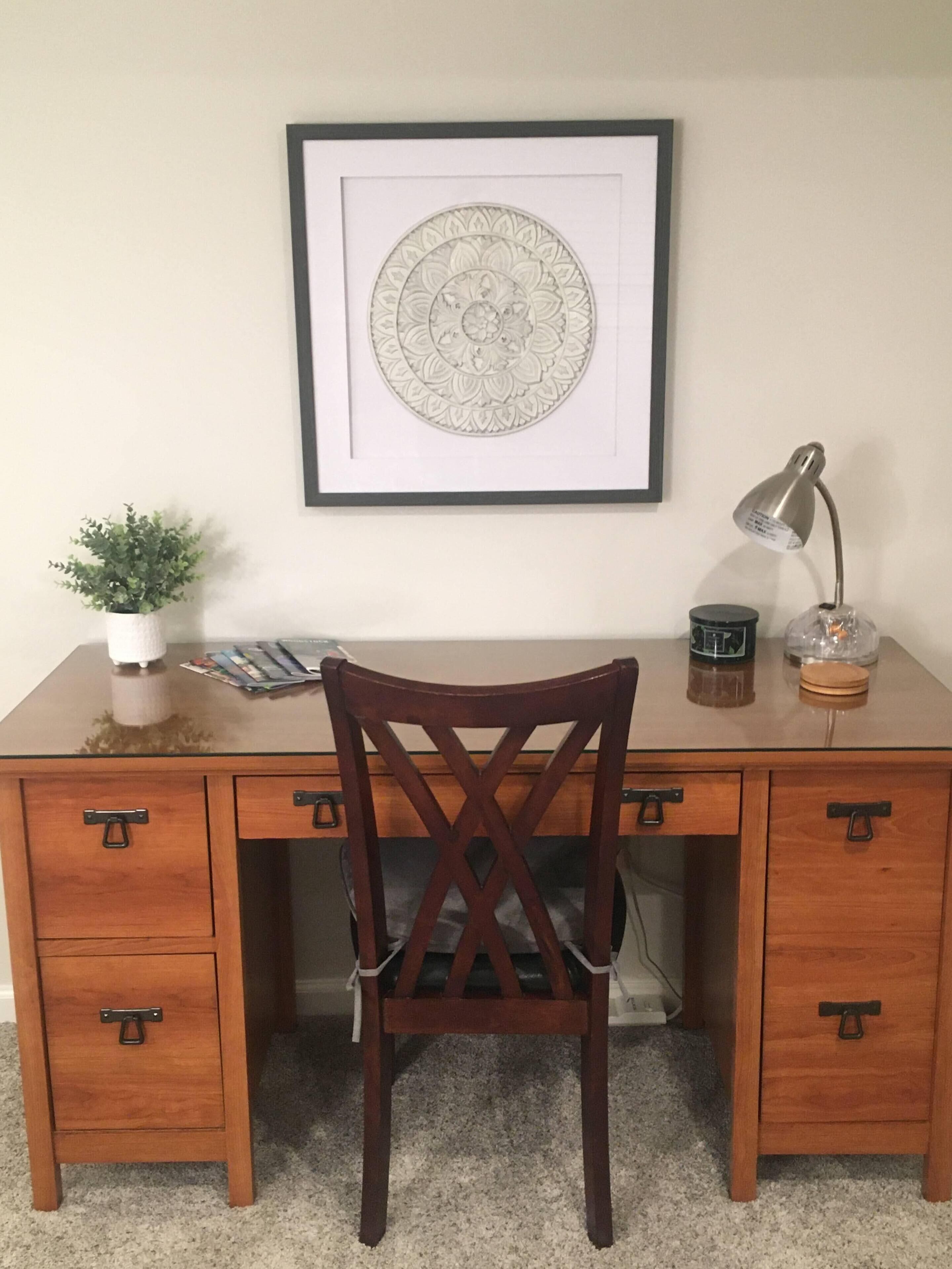 Een houten bureau met een donkerhouten stoel, een lijst met een afbeelding erboven, een kleine potplant, een lamp en een paar voorwerpen op het bureau.
