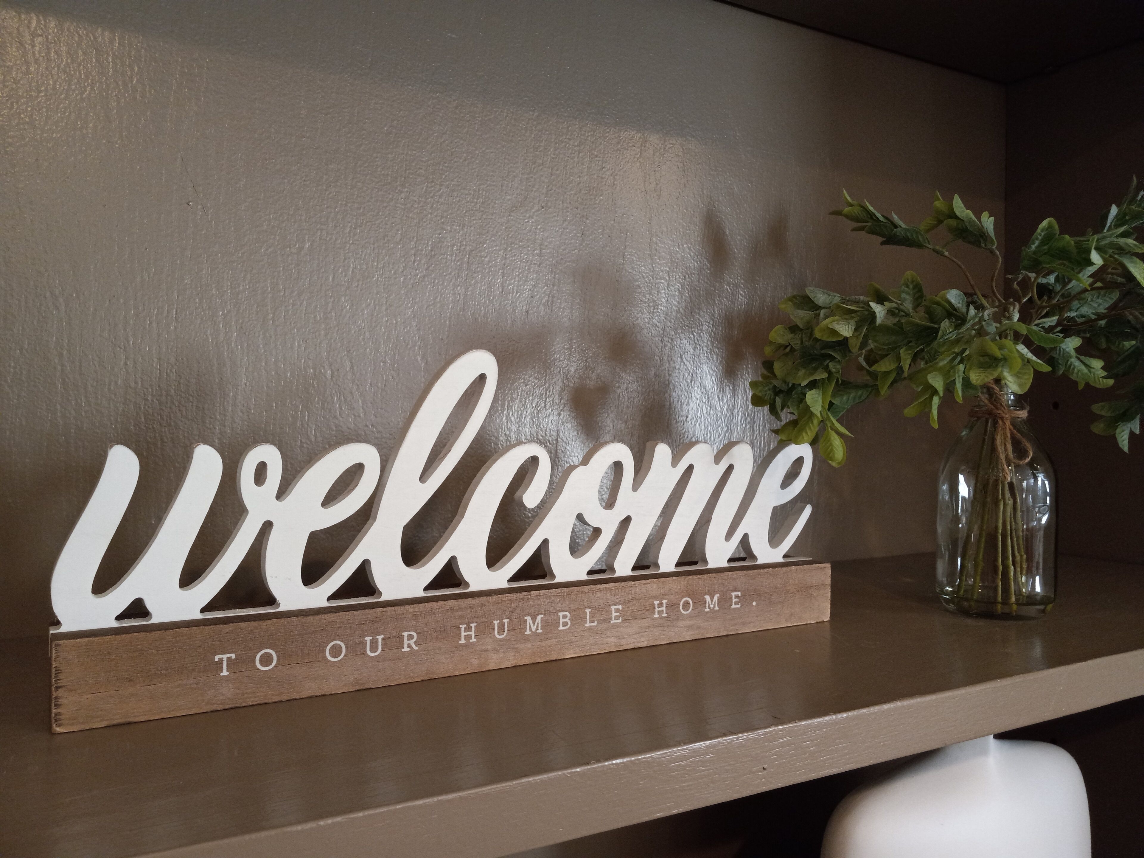 A wooden sign with the words "welcome to our humble home" on a shelf.