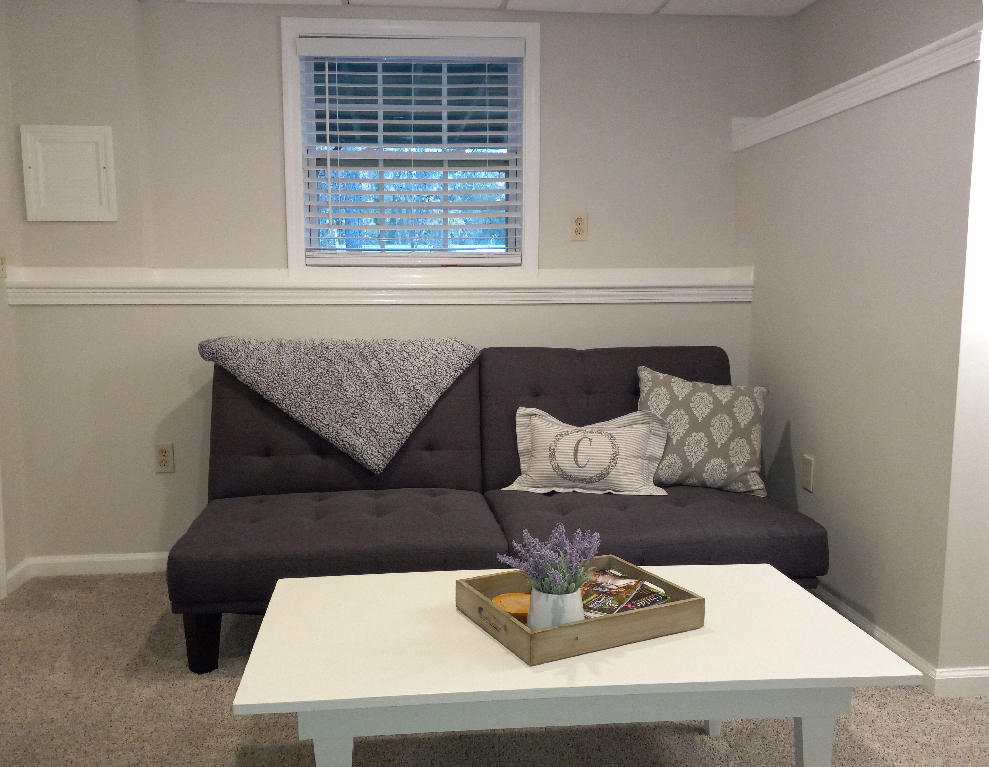 A living room with a dark grey sofa, white cushions, a white coffee table, and a window with blinds.