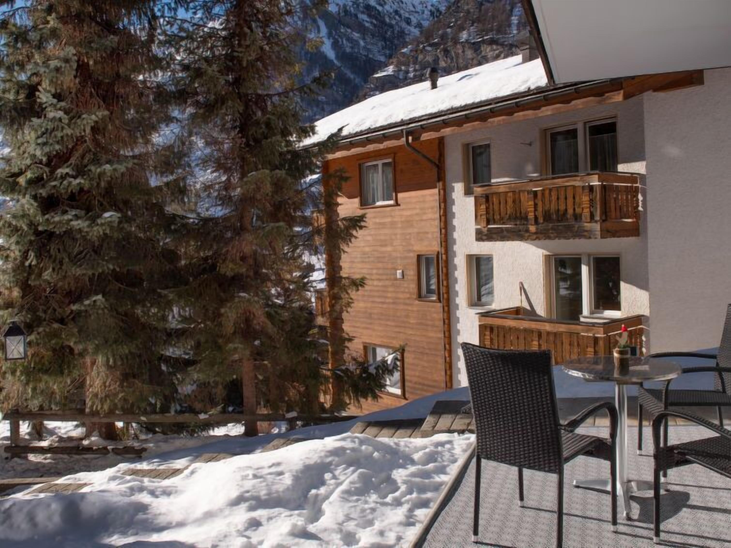Een besneeuwde buitenruimte met een houten terras, een tafel met stoelen en een gebouw met een houten balkon.
