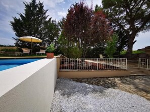 Une piscine entourée d’une clôture blanche, une terrasse en pierre et un arbre aux feuilles rouges.