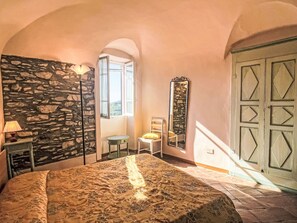 A bedroom with a stone wall, a bed with a patterned bedspread, a nightstand with a lamp, a small table with a chair, and a mirror on the wall.