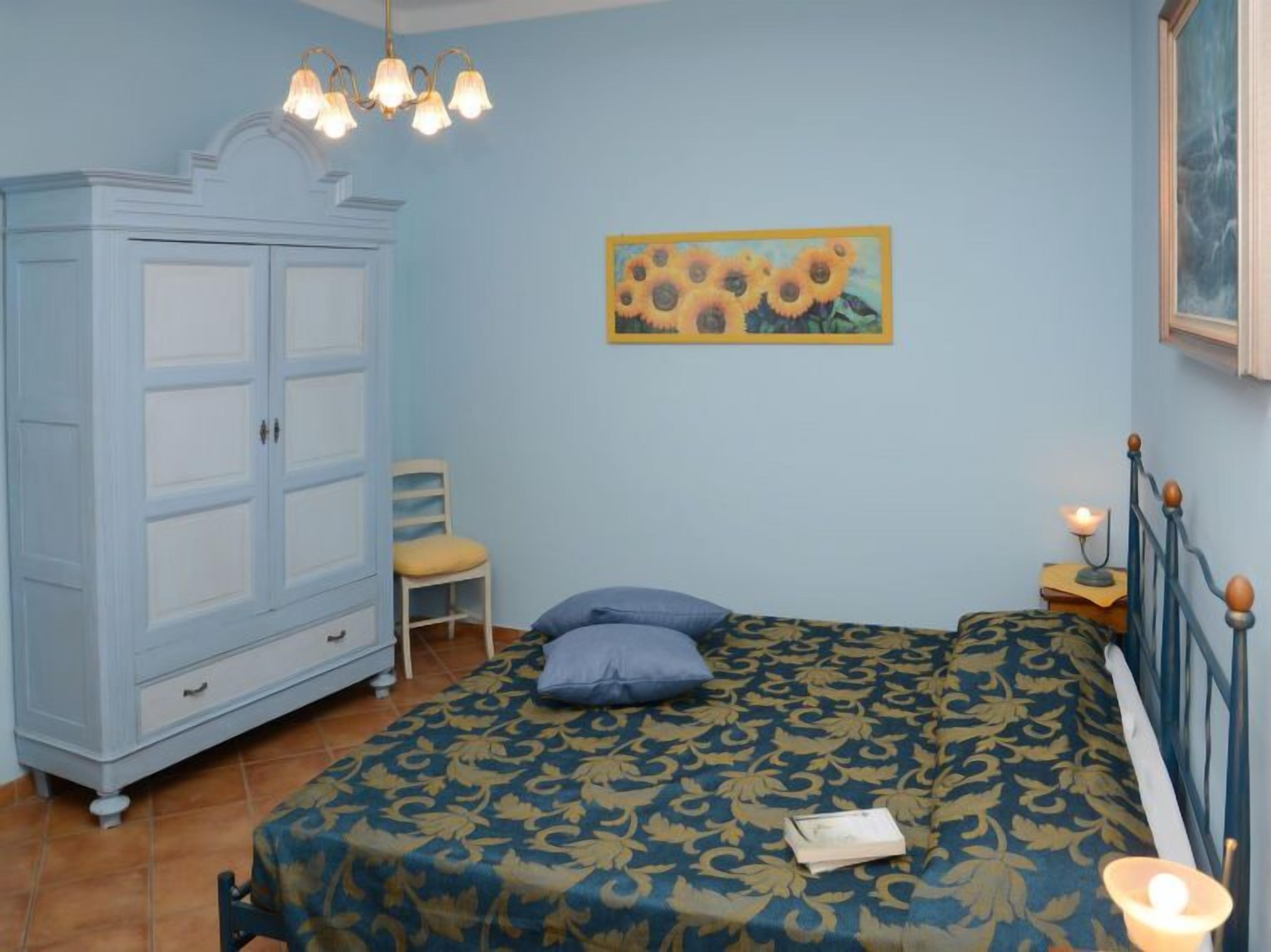 A bedroom with a bed, a wardrobe, a chair, a painting, and a nightstand with a lamp.