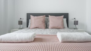 Un letto rifatto con una coperta a fantasia, due cuscini e due asciugamani piegati su un tappetino rosa ruvido.