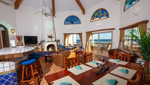 Uma sala de estar ampla, com uma grande mesa de madeira, banquetas de bar e uma lareira. Várias janelas oferecem vista para o oceano.