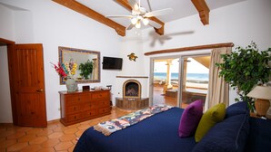 Quarto com cama, cômoda, televisão, lareira e vista para a praia através de uma porta de vidro.