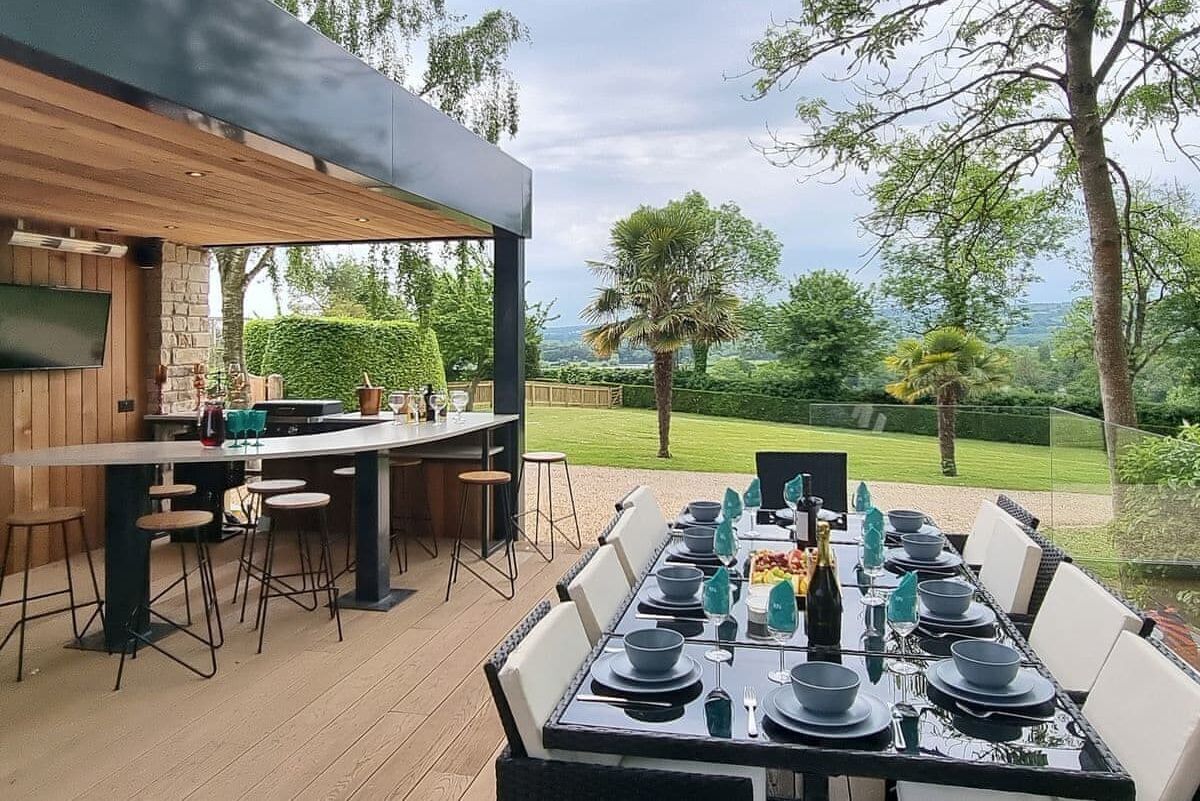 An outdoor dining area with a long table set for six, surrounded by bar stools. A wooden pergola provides shade, and a TV is mounted on the wall.