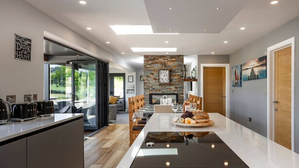 A modern kitchen with a central island, a dining area with a stone wall, and a view through a glass door. - Kingsley Lake View & Paddocks - A group retreat with hot tubs, sports bars & spectacular lake views (Chew Stoke)