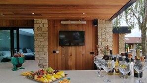 A bar area with a stone wall, wooden ceiling, and a flat-screen TV. There are glasses, bottles, and a fruit platter on the counter. - Kingsley Lake View & Paddocks - A group retreat with hot tubs, sports bars & spectacular lake views (Chew Stoke)