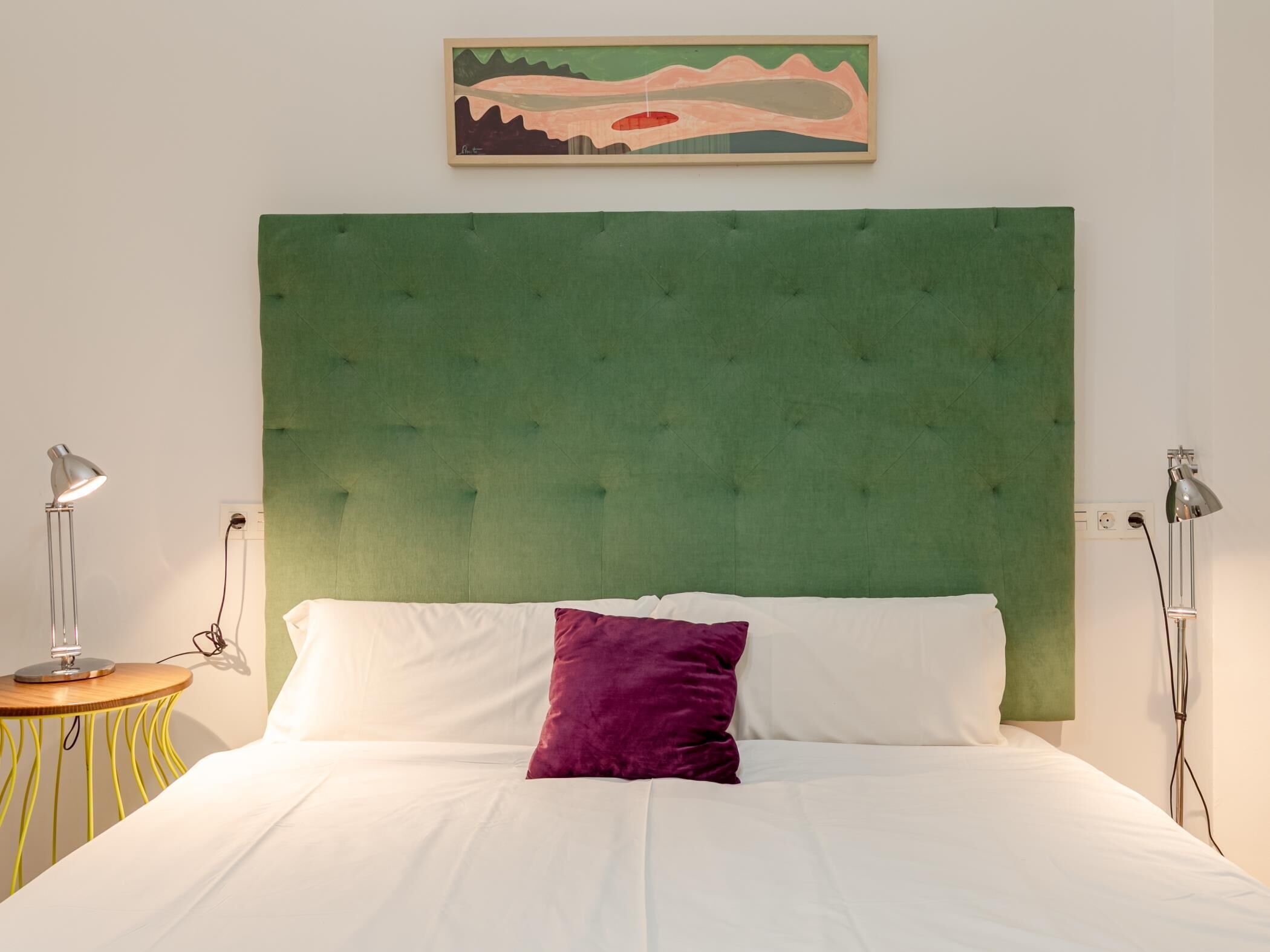 Un lit avec une tête de lit recouverte d’un tissu vert, une literie blanche et un oreiller violet. Deux lampes de chevet sont placées de chaque côté du lit.