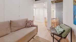 A modern living room with a beige sofa, patterned cushions, a small wooden table with chairs, and a large window. - Plaza del Pan Deluxe 2 (Sevilla)