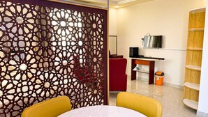 A modern room with a decorative partition, a round table with yellow chairs, a red chair, a desk with a computer, a TV, and a wooden shelving unit. - Discovery Guest House -Sur (Sur)