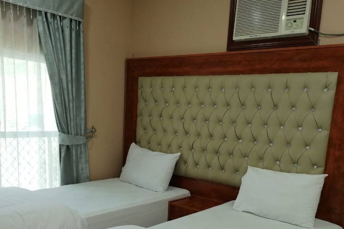 A hotel room with two single beds, a tufted headboard, a window with curtains, and an air conditioning unit above the bed.