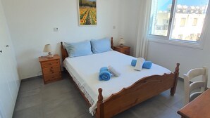 A bedroom with a wooden bed, a nightstand, a lamp, a chair, and a window with a view of buildings. - Ithaki 2 Bedroom Penthouse (Λατσί/ Πόλη Χρυσοχούς)