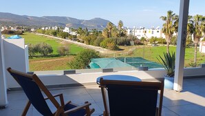 A covered outdoor seating area with two wooden chairs and a table, overlooking a green landscape with buildings and mountains in the distance. - Ithaki 2 Bedroom Penthouse (Λατσί/ Πόλη Χρυσοχούς)