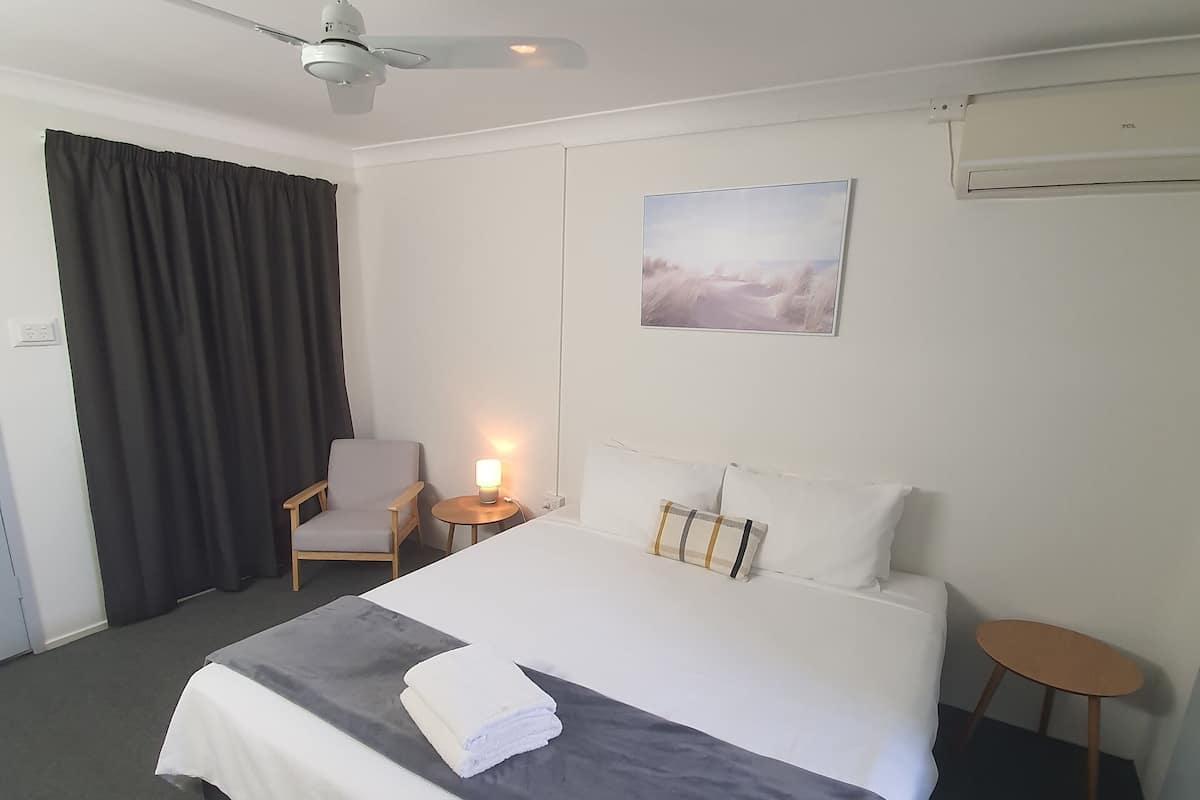 A hotel room with a bed, a chair, a small table, a ceiling fan, and a wall-mounted air conditioner.