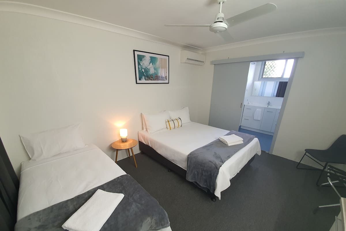 A hotel room with two beds, a ceiling fan, a small table with a lamp, a painting on the wall, and a view of the outside through a window.