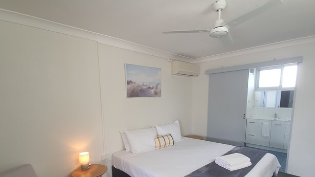A hotel room with a bed, a chair, a small table with a lamp, an air conditioning unit, and a window. - Sun City Motel (Bundaberg North)