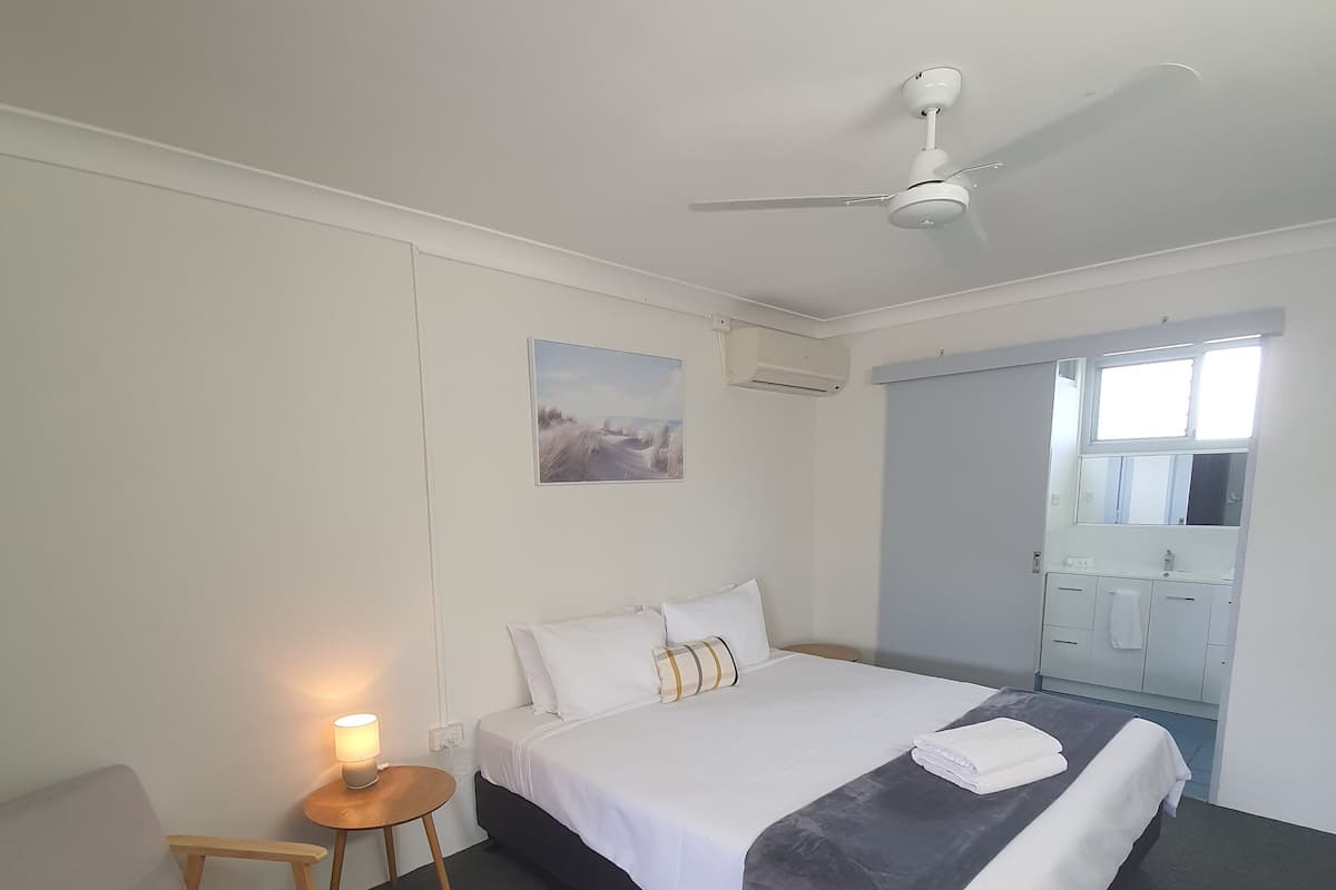 A hotel room with a bed, a chair, a small table with a lamp, an air conditioning unit, and a window.