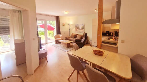 En moderne stue med sofa, sofabord og spisebord. Der er et køkkenområde med komfur og skabe. Rummet har en skydedør af glas, der fører til en terrasse med parasol.