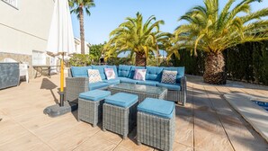 Un espace extérieur aménagé avec un canapé d’angle, des poufs et une table, le tout entouré de palmiers et d’une piscine.