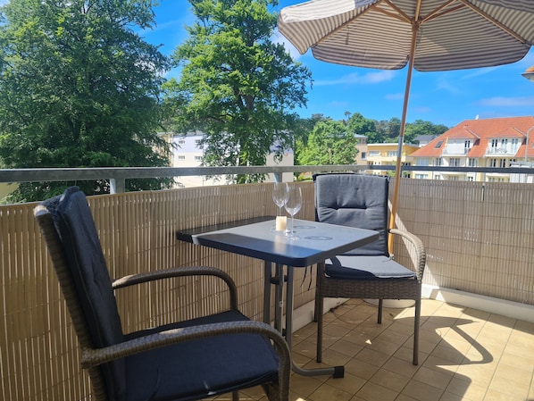 A balcony with a table, two chairs, an umbrella, and a view of buildings and trees.