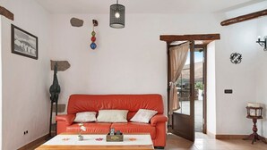 Un salon avec un canapé rouge, une table basse en bois, un tableau encadré, un luminaire suspendu et une porte donnant sur l’extérieur.