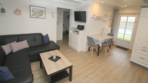 Una sala de estar moderna con un sofá seccional negro, una mesa de centro de madera, una zona de comedor con una mesa pequeña y sillas, un televisor montado en la pared y un armario blanco.